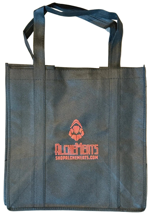 Black AlcheMeats tote bag with red logo and text on a wooden floor