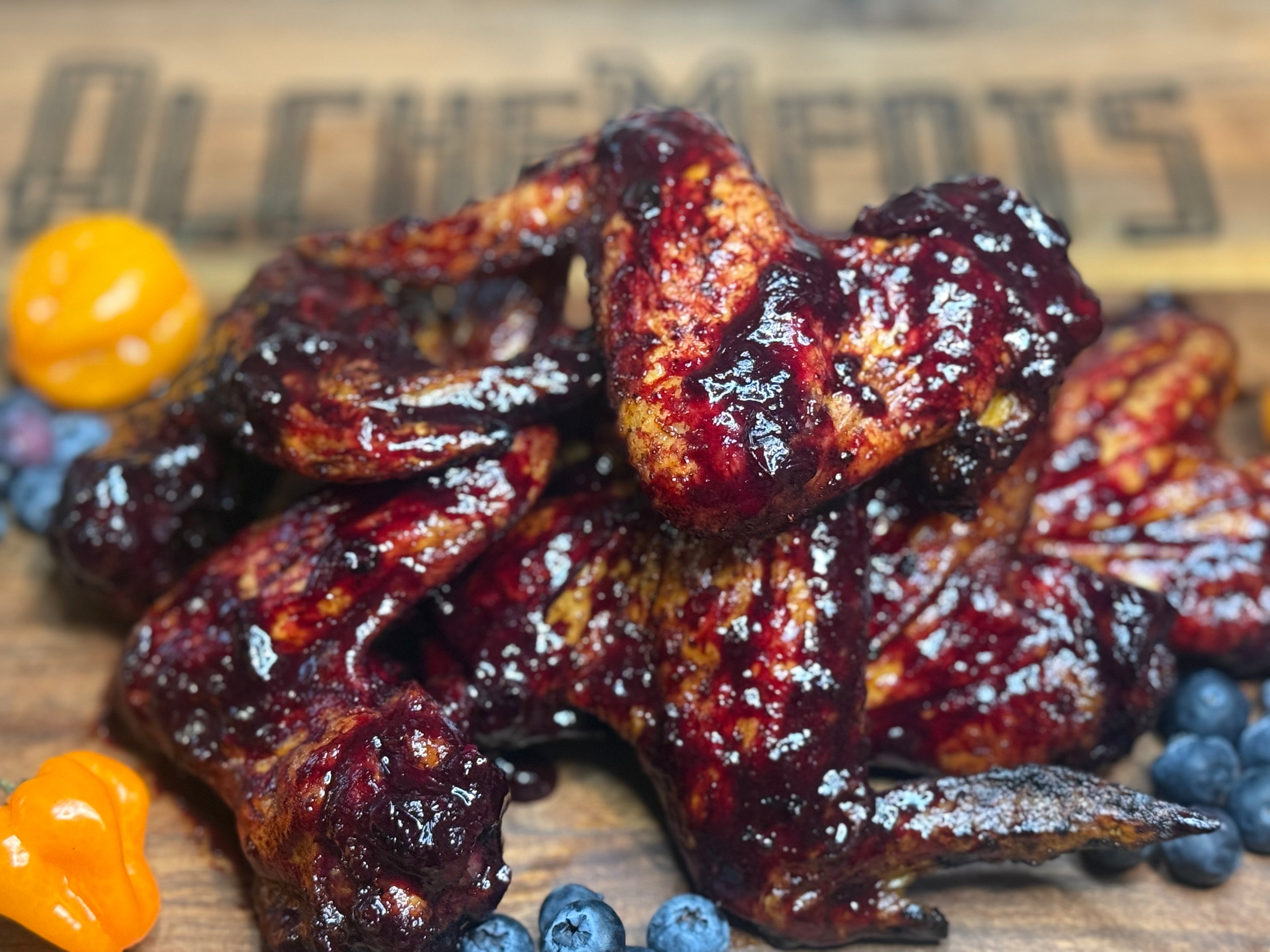 Spicy wings with blueberries and orange peppers on a wooden board with 'AlcheMeats' branding.